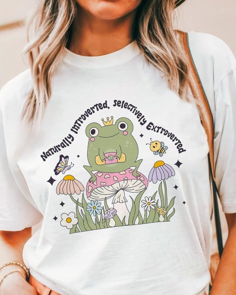 White t-shirt featuring a cartoon frog wearing a crown and sitting on a mushroom, surrounded by flowers and a butterfly; text reads Naturally Introverted, Selectively Extroverted.