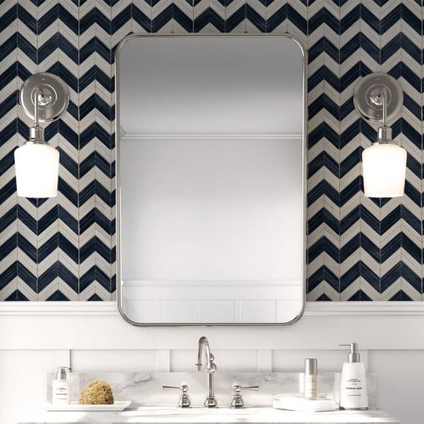 Bathroom Vanity With A Rectangular Mirror Flanked By Wall-Mounted Lights, All Set Against A Herringbone Patterned Tile Backsplash.