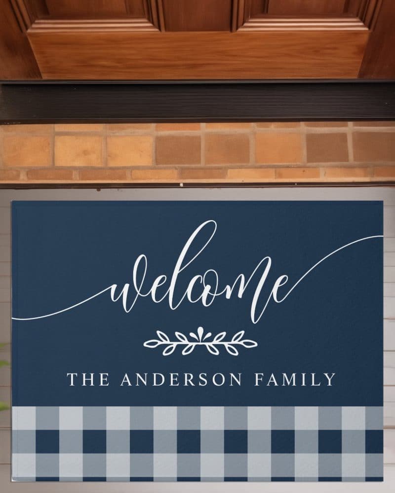 Navy blue welcome mat with white script lettering that says Welcome, The Anderson Family, and a bottom border of blue and gray gingham.
