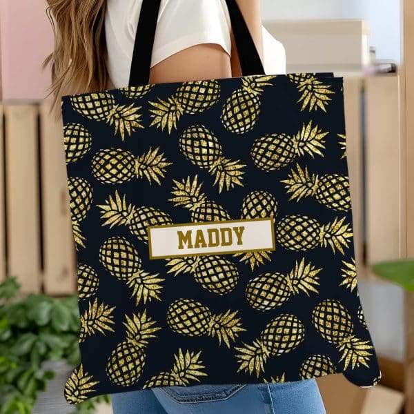 A Black Tote Bag With A Gold Glitter Pineapple Pattern And The Name Maddy.