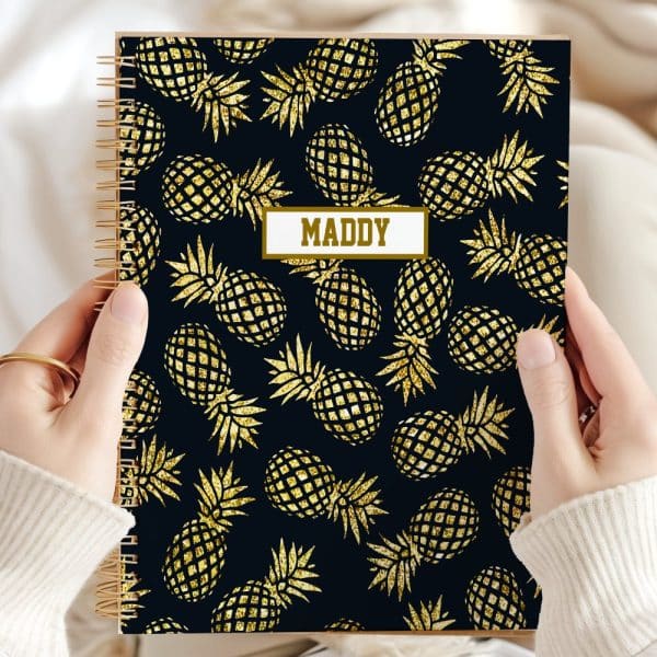 Two Hands Hold A Spiral Notebook With A Dark Background And A Gold Glitter Pineapple Pattern. The Name Maddy Is Printed On The Cover.