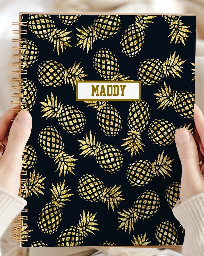 Two hands hold a spiral notebook with a dark background and a gold glitter pineapple pattern. The name Maddy is printed on the cover.