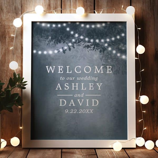White Framed Wedding Welcome Sign With Ashley And David'S Names And Wedding Date. The Background Features A Dark Blue Design With String Lights.