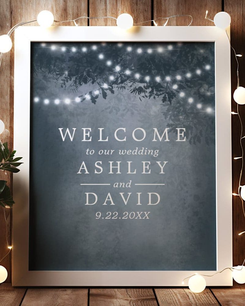 White framed wedding welcome sign with Ashley and David's names and wedding date. The background features a dark blue design with string lights.