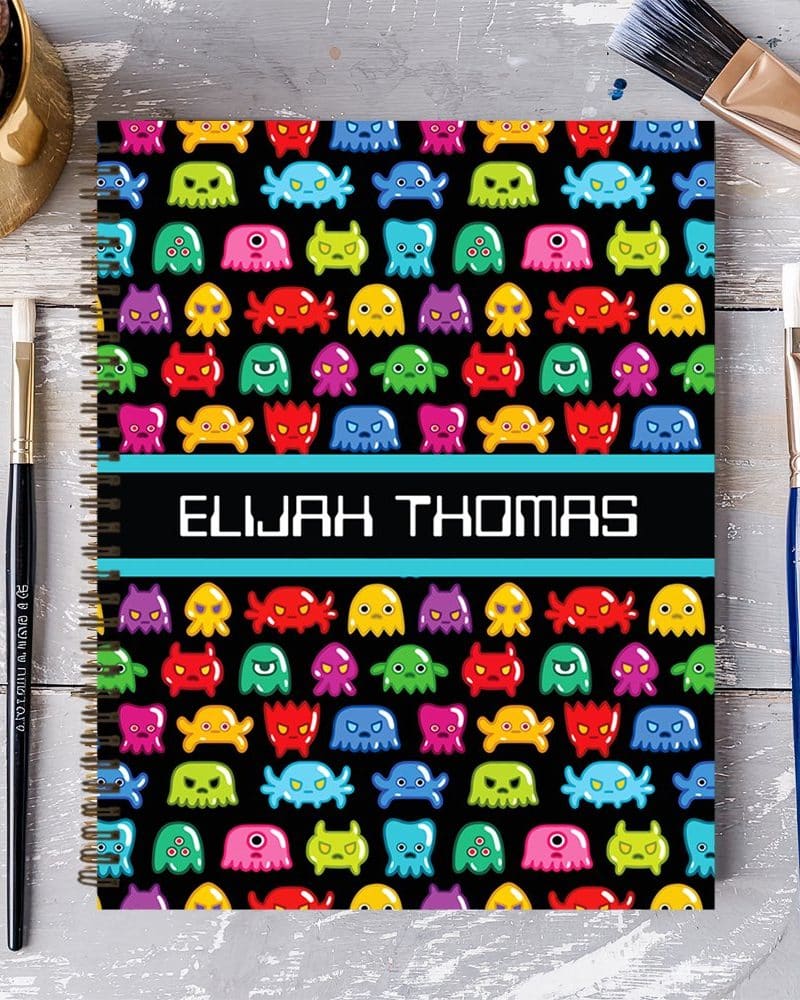 Spiral notebook with a black cover featuring a repeating pattern of colorful cartoon monsters and the name Elijah Thomas.
