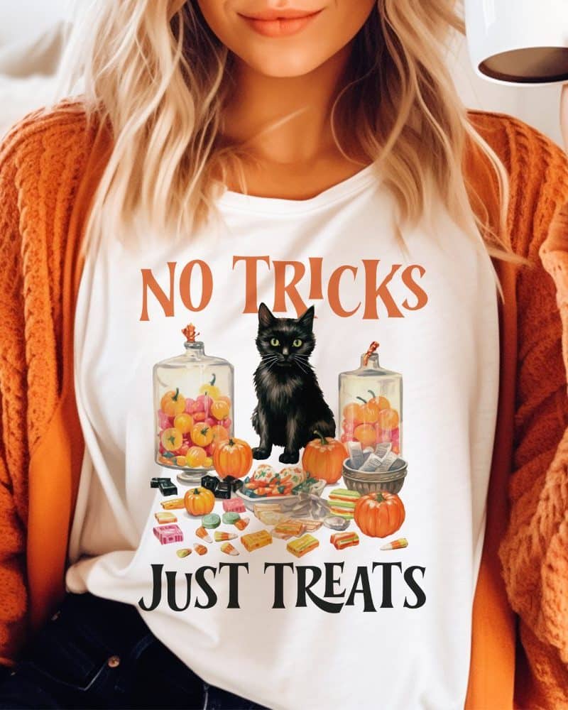 White shirt with a black cat sitting amidst Halloween candy and pumpkins. The text "No Tricks Just Treats" is printed on the shirt.