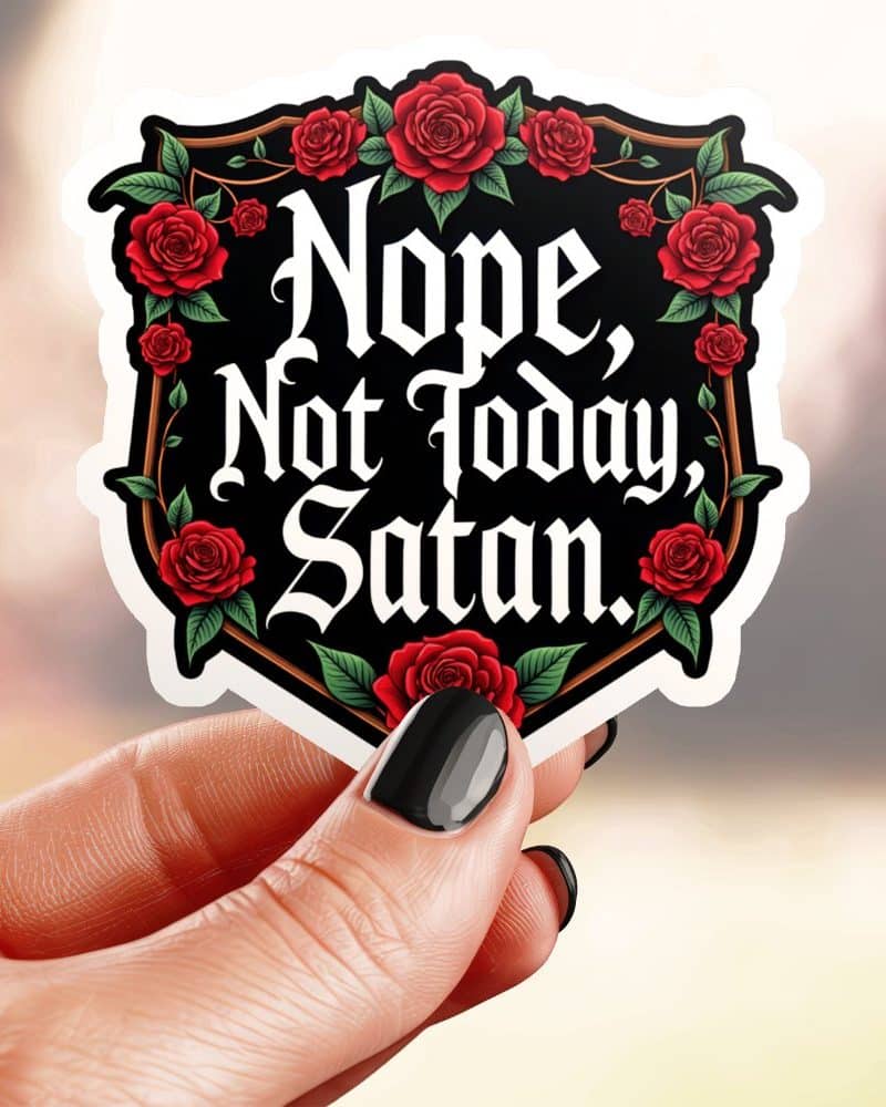 A hand holds a sticker with a black shield shape bordered with red roses and the text Nope, Not Today, Satan in a gothic style font.
