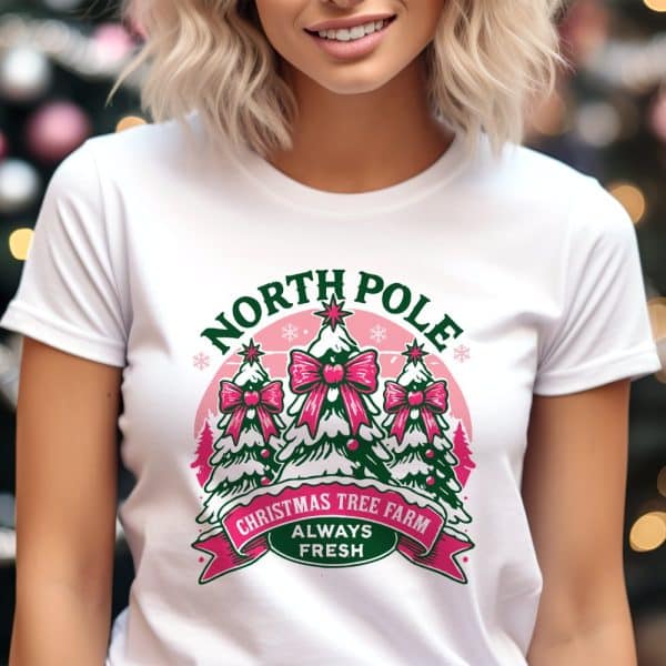 Woman Wearing A White T-Shirt With A Christmas Tree Farm Design. The Design Features Three Christmas Trees With Pink Bows, The Words North Pole And Always Fresh.