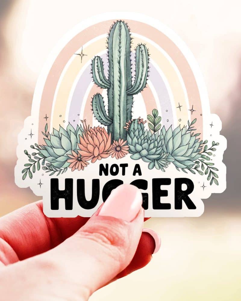 Hand holding a sticker featuring a cactus and succulents with a rainbow arc behind them, and the text Not a Hugger.