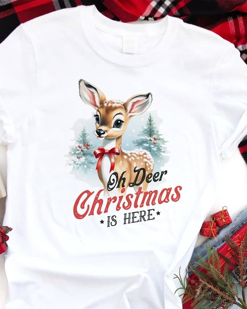 White t-shirt featuring a vintage-style illustration of a fawn wearing a red bow, surrounded by a snowy winter scene. Text reads Oh Deer Christmas Is Here.