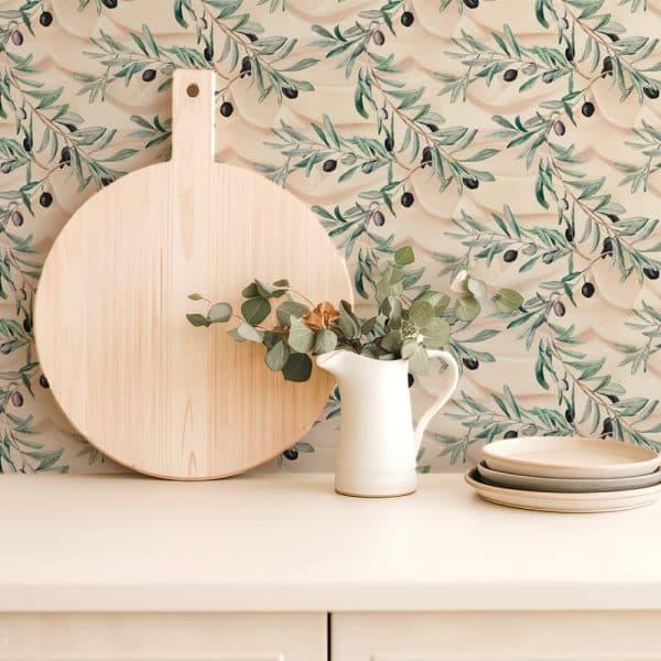 Large Wooden Cutting Board, White Pitcher With Eucalyptus, And Stacked Plates Sit On A Counter Against A Backdrop Of Olive Branch Wallpaper.