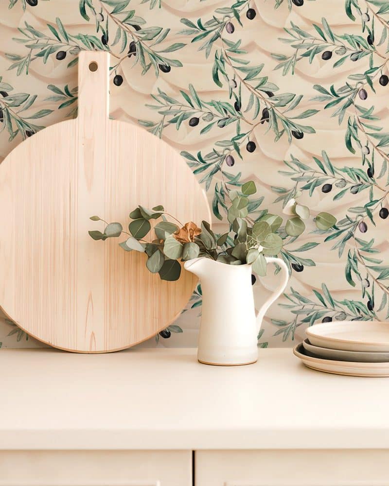 Large wooden cutting board, white pitcher with eucalyptus, and stacked plates sit on a counter against a backdrop of olive branch wallpaper.
