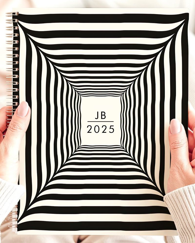 Hands holding a 2025 spiral-bound notebook with a black and white striped, tunnel-like design and the initials JB.