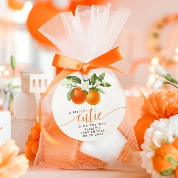 A Sheer Orange And White Favor Bag With An Orange Blossom And Citrus Fruit Design, Tied With An Orange Ribbon, And A Round Tag That Reads A Little Cutie Is On The Way, Everly'S Baby Shower, 08.20.2033.