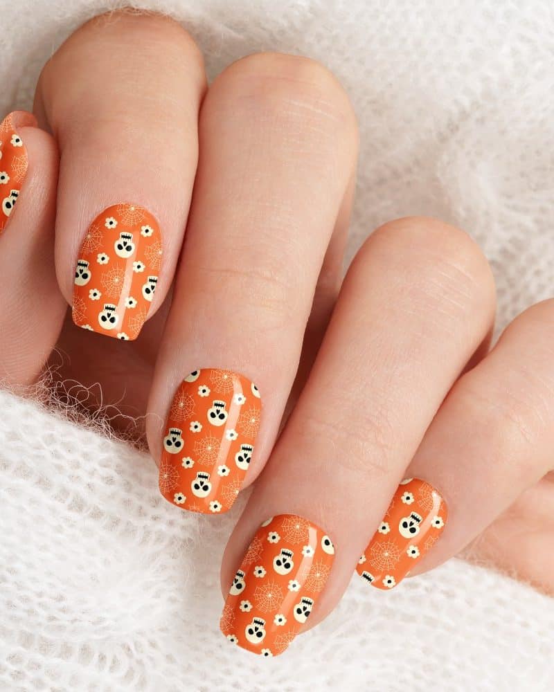 Close-up of a hand with Halloween-themed nail art. Orange nails are decorated with a pattern of small skulls and spiderwebs.