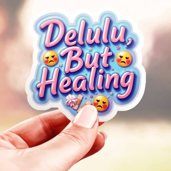 Hand Holding A Sticker That Says Delulu, But Healing In Pink Lettering On A Light Blue Background, With Sad Emojis And Bandages.