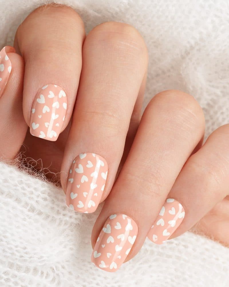 Close-up of a hand with peach-colored nails decorated with small white hearts.