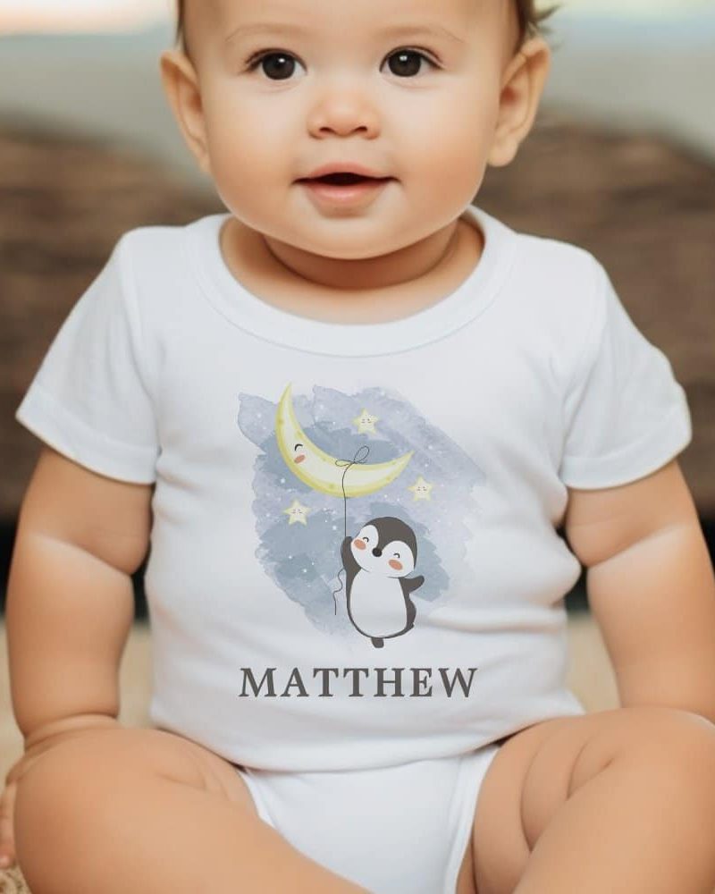 Baby wearing a white onesie with a penguin holding a crescent moon. The onesie is personalized with the name Matthew.