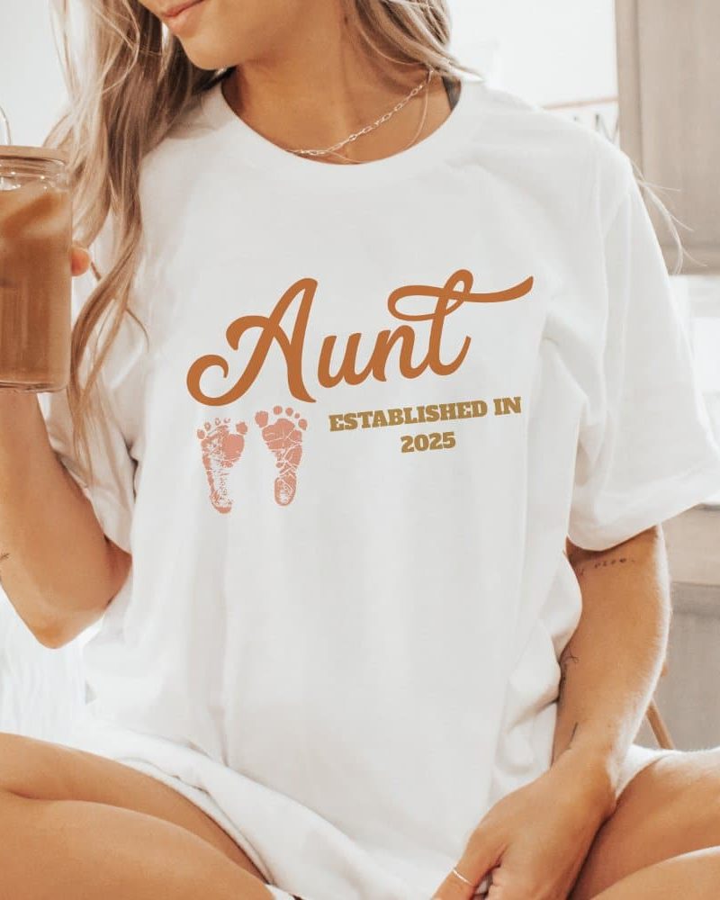 Woman wearing a white t-shirt that says Aunt established in 2025 with baby footprints.
