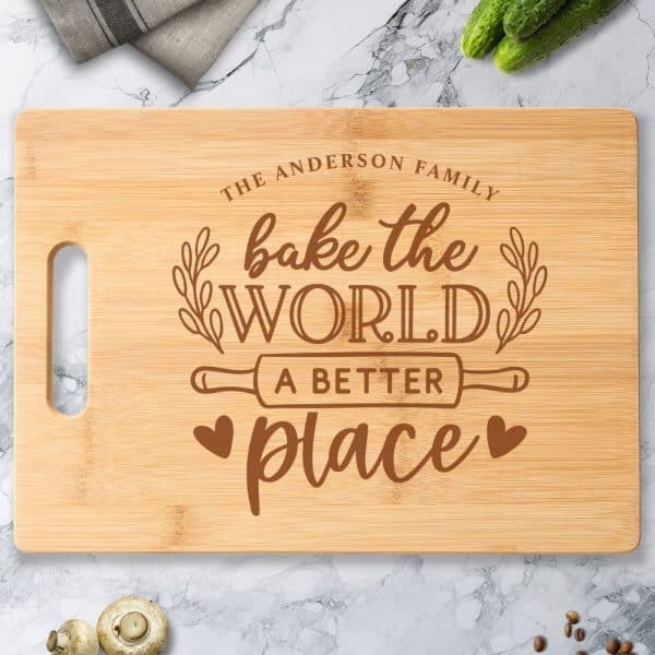 Light Brown Bamboo Cutting Board With A Handle, Engraved With The Text Bake The World A Better Place, Along With The Words The Anderson Family.