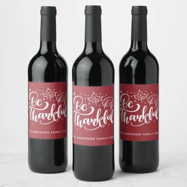 Three Wine Bottles With Maroon Labels That Read Be Thankful In White Script Lettering, Accented With Small White Leaf Drawings.