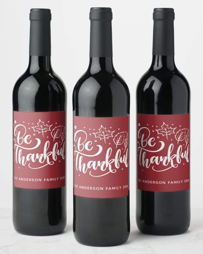 Three wine bottles with maroon labels that read Be Thankful in white script lettering, accented with small white leaf drawings.