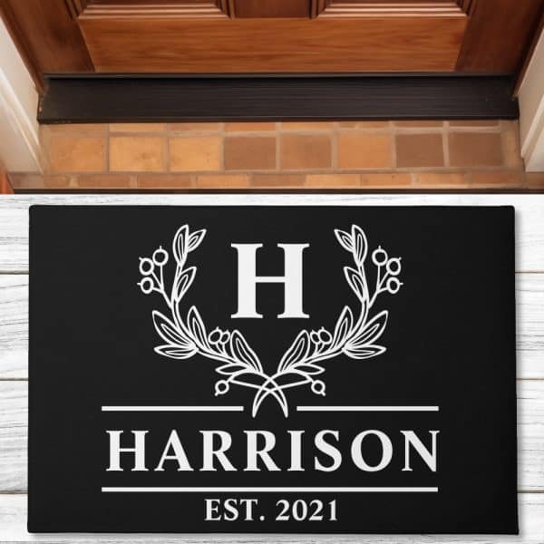 Black Doormat With A White Monogram: The Letter H, Flanked By Leaf Sprigs, With Harrison And Est. 2021 Below.