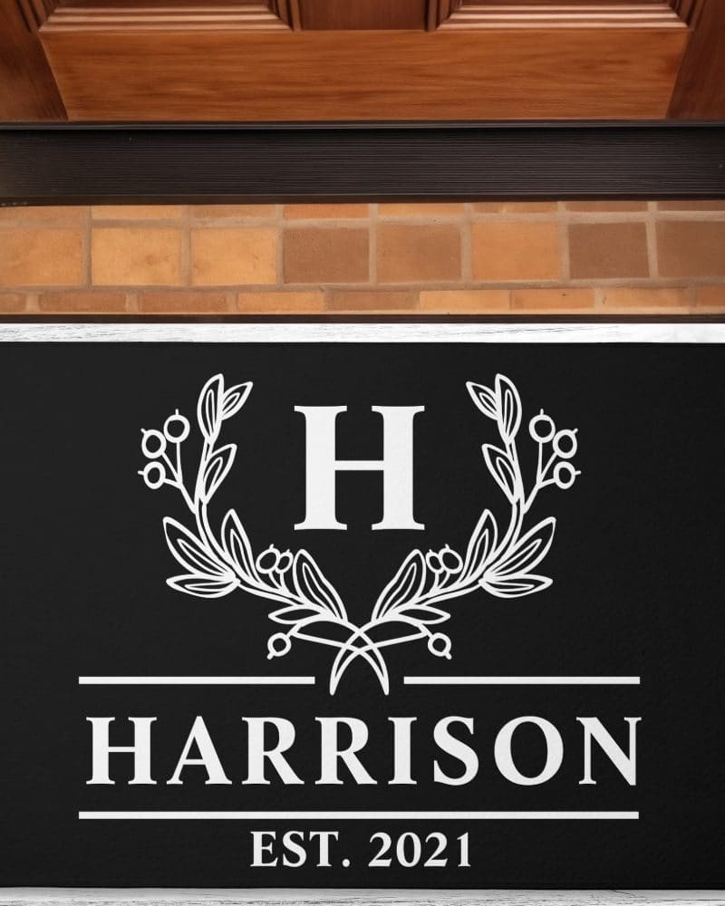 Black doormat with a white monogram: the letter H, flanked by leaf sprigs, with Harrison and Est. 2021 below.