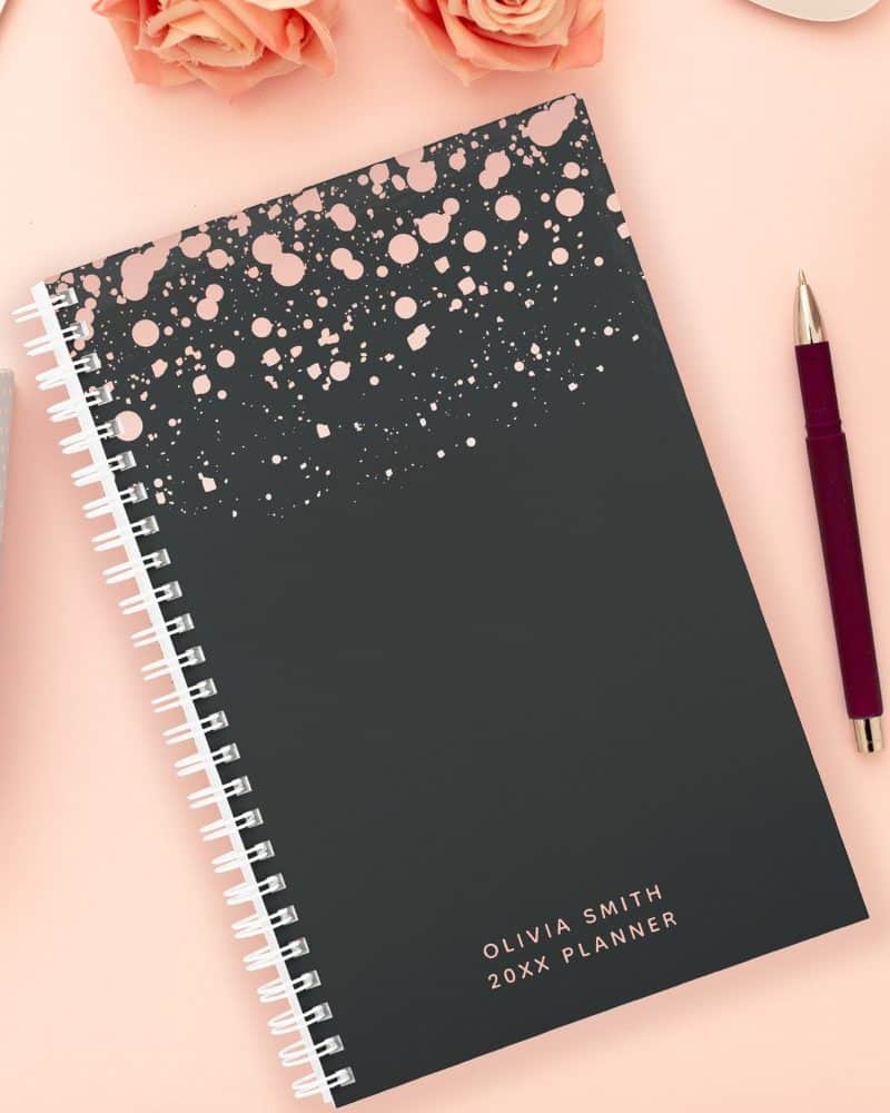 Dark gray spiral-bound planner with rose gold splatter design and personalized name.