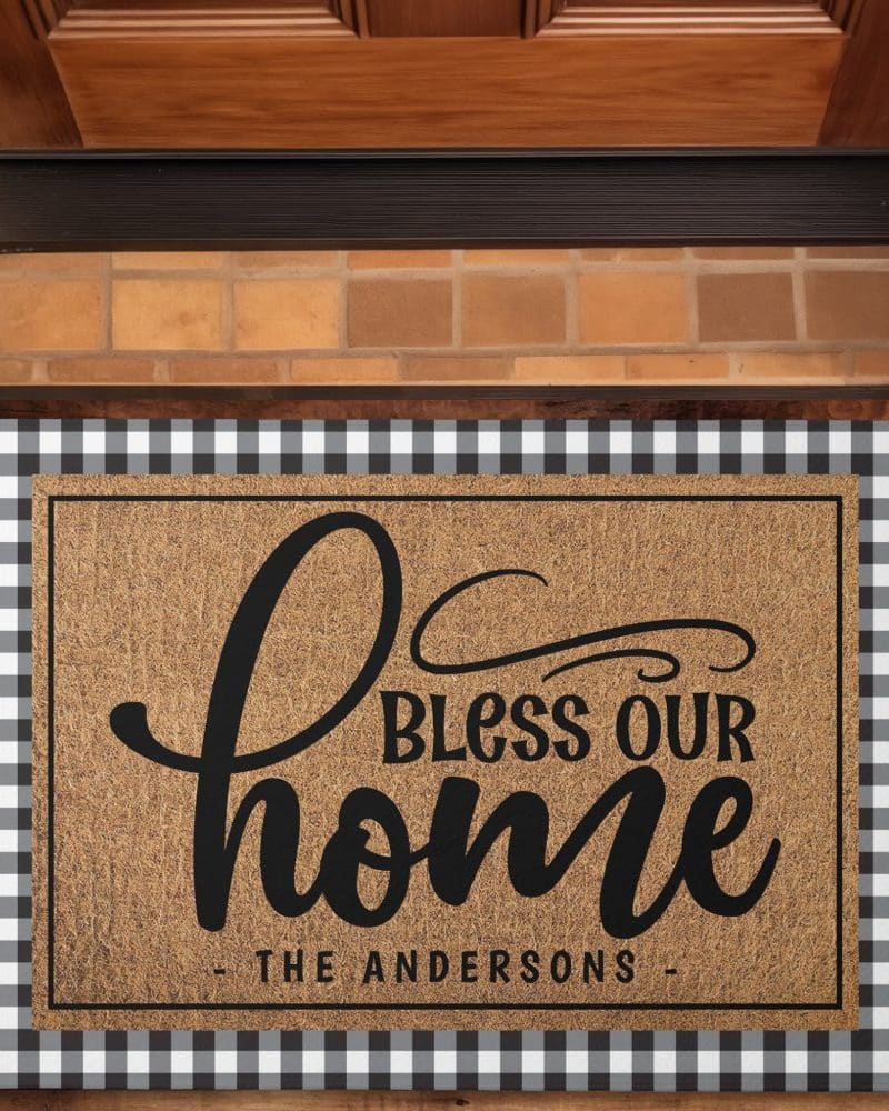 Brown coir doormat with black lettering that says Bless Our Home and the Andersons name below. The doormat has a black and white checkered border.