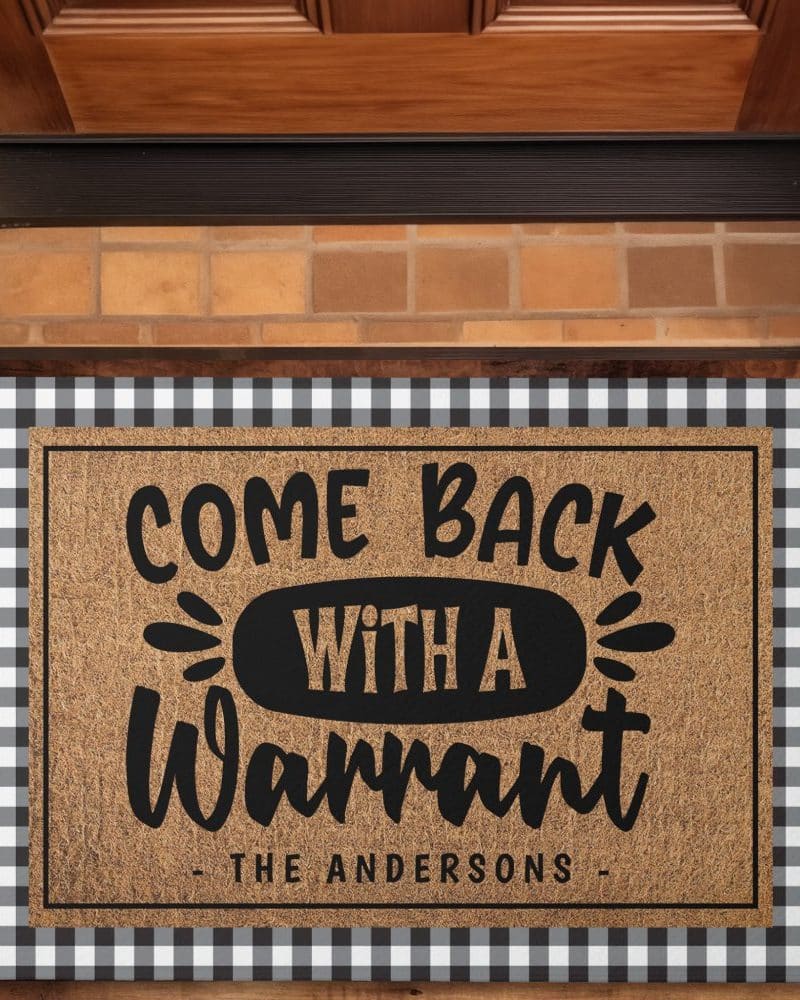 A coir doormat with black lettering that says Come Back With A Warrant, and The Andersons at the bottom. The doormat is on a black and white checkered border.