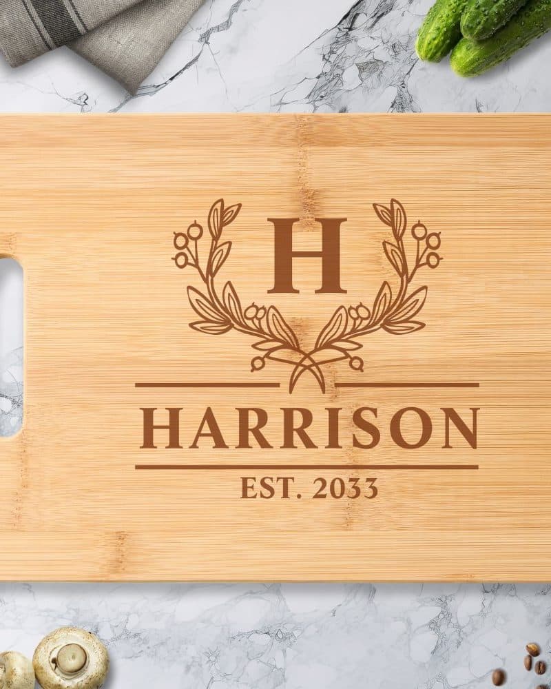 Light brown bamboo cutting board with a monogrammed design. The design includes the initial H, the last name Harrison, and the date 2033.