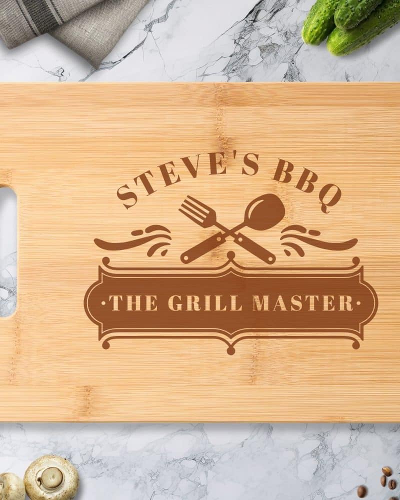 Personalized bamboo cutting board with Steves BBQ, The Grill Master engraved in brown lettering, and crossed fork and spoon graphic.
