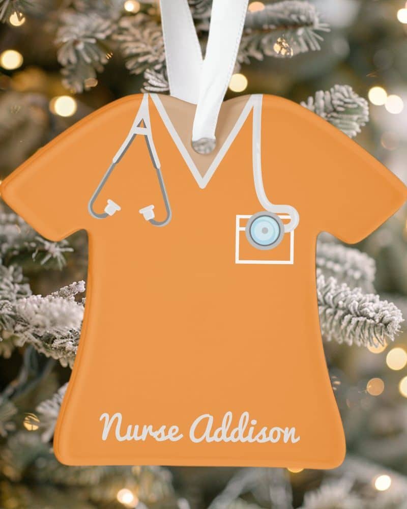 Orange Christmas ornament shaped like a nurse's scrub top, with a stethoscope and the name Nurse Addison.