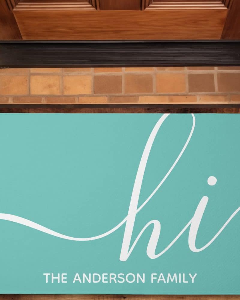 Teal welcome mat with the word hi in a stylized white script font and The Anderson Family printed below.
