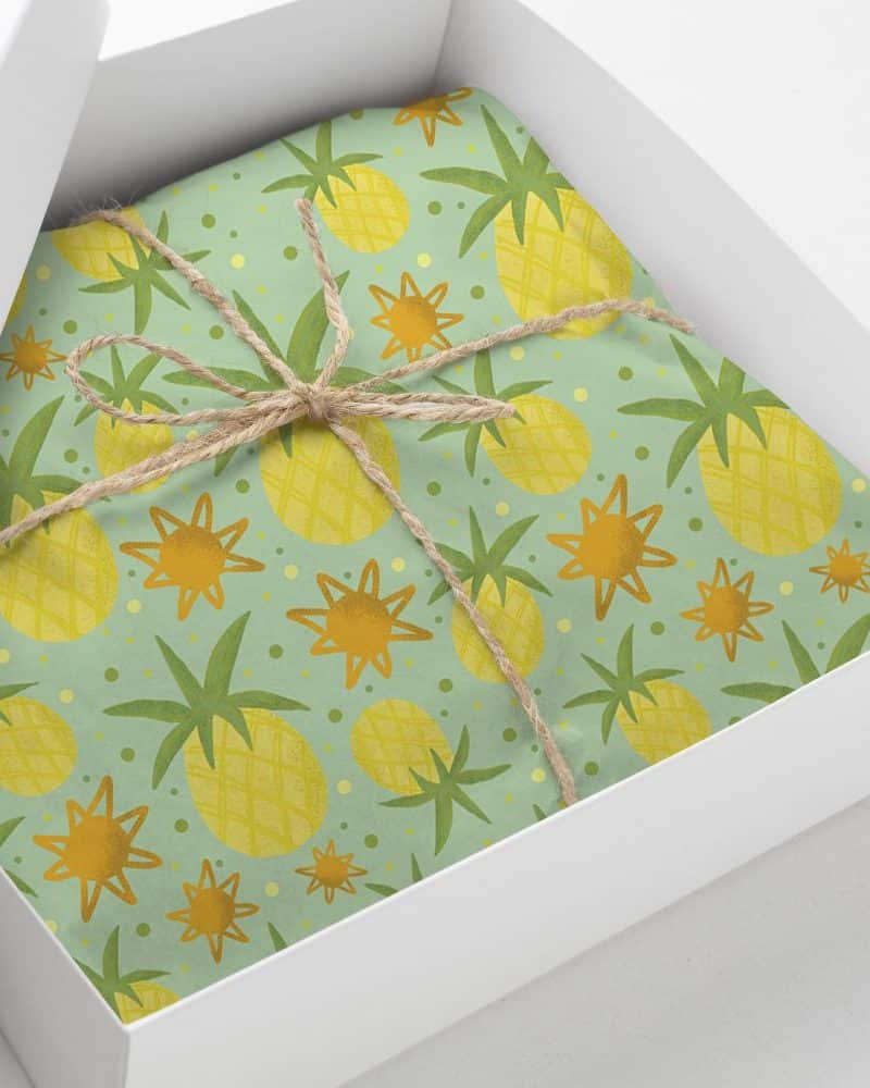 Gift box with light green wrapping paper featuring a repeating pattern of yellow pineapples and orange stars. The box is tied with twine.