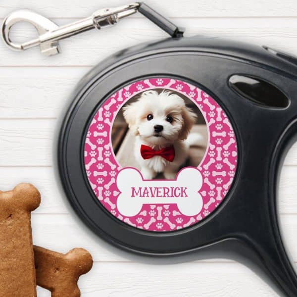 A Retractable Dog Leash With A Pink Bone-Patterned Circle Featuring A Photo Of A Small White Dog Wearing A Red Bow Tie And The Name Maverick.