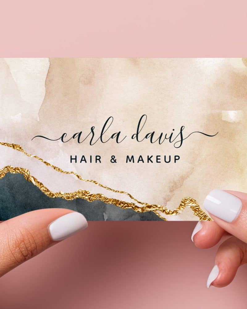 Two hands hold up a business card. The card features a watercolor design in beige and dark blue with gold accents, and the name Carla Davis Hair & Makeup.