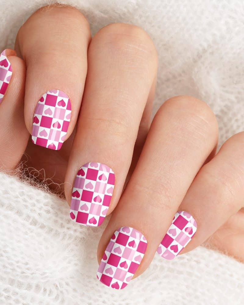 Hand with fingernails painted with a pink and white checkered pattern featuring small pink hearts.