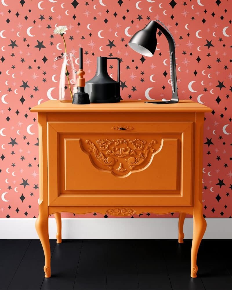 Orange painted wood cabinet with ornate detailing, holding a lamp and decorative objects.