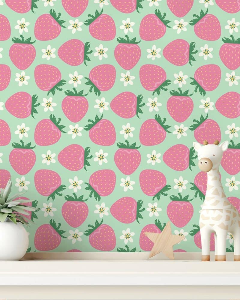 A mint green wall covered in a repeating pattern of pink strawberries and white flowers. A small giraffe toy and a plant are in front of the wall.