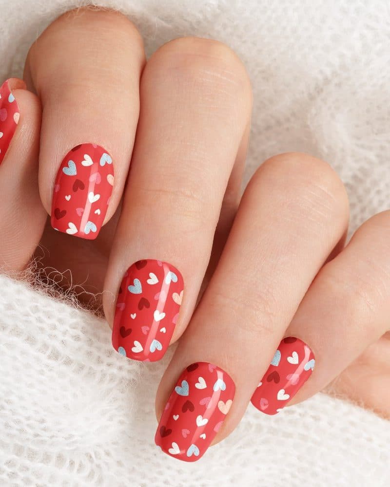 Close-up of a hand with red nails decorated with small white, pink, and light blue hearts.