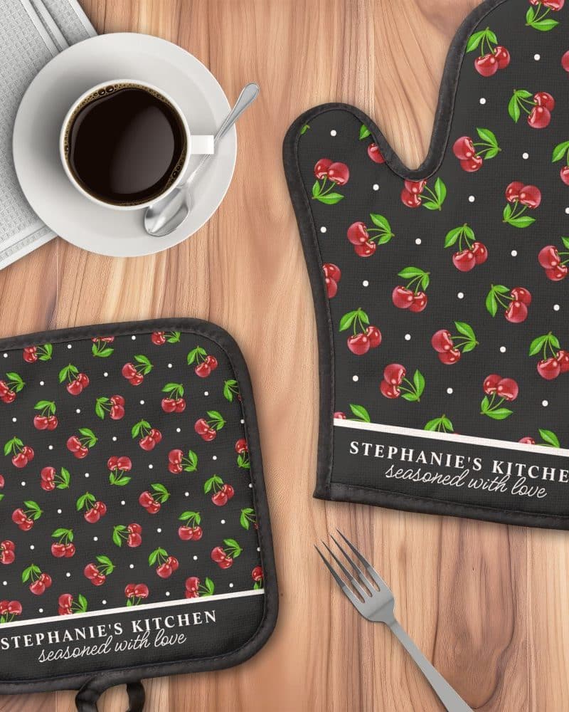 A black oven mitt and pot holder with a red cherry pattern and personalized text.