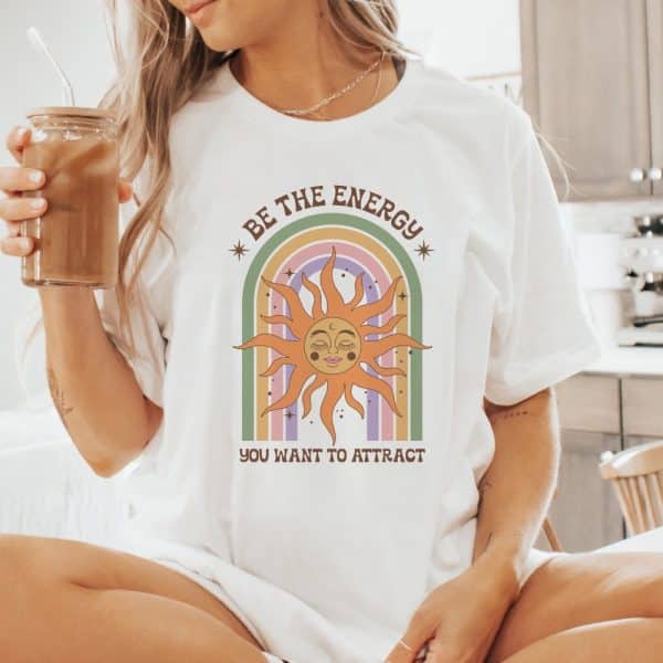 White T-Shirt Featuring A Sun With A Face In A Rainbow. Text Reads, Be The Energy You Want To Attract.