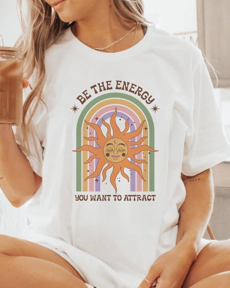 White t-shirt featuring a sun with a face in a rainbow. Text reads, Be the energy you want to attract.