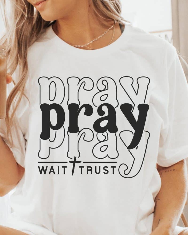 Woman wearing a white t-shirt with the word pray stacked three times in a retro design, and the words wait trust beneath it.