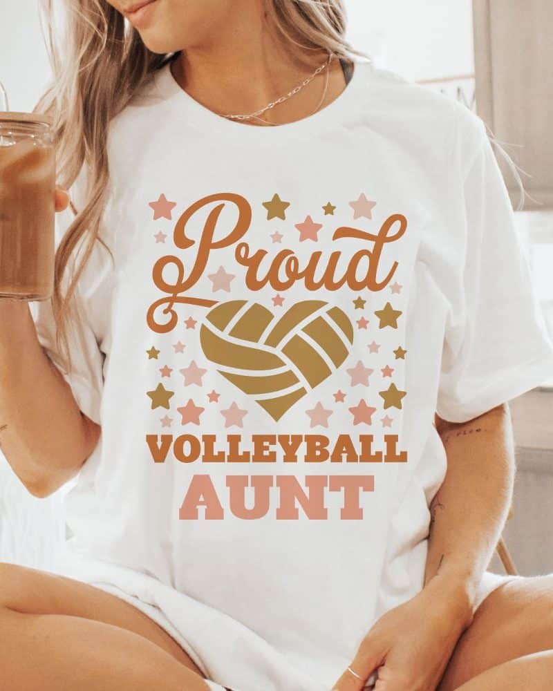 A woman wearing a white t-shirt that says Proud Volleyball Aunt in gold and pink lettering with a volleyball heart design.