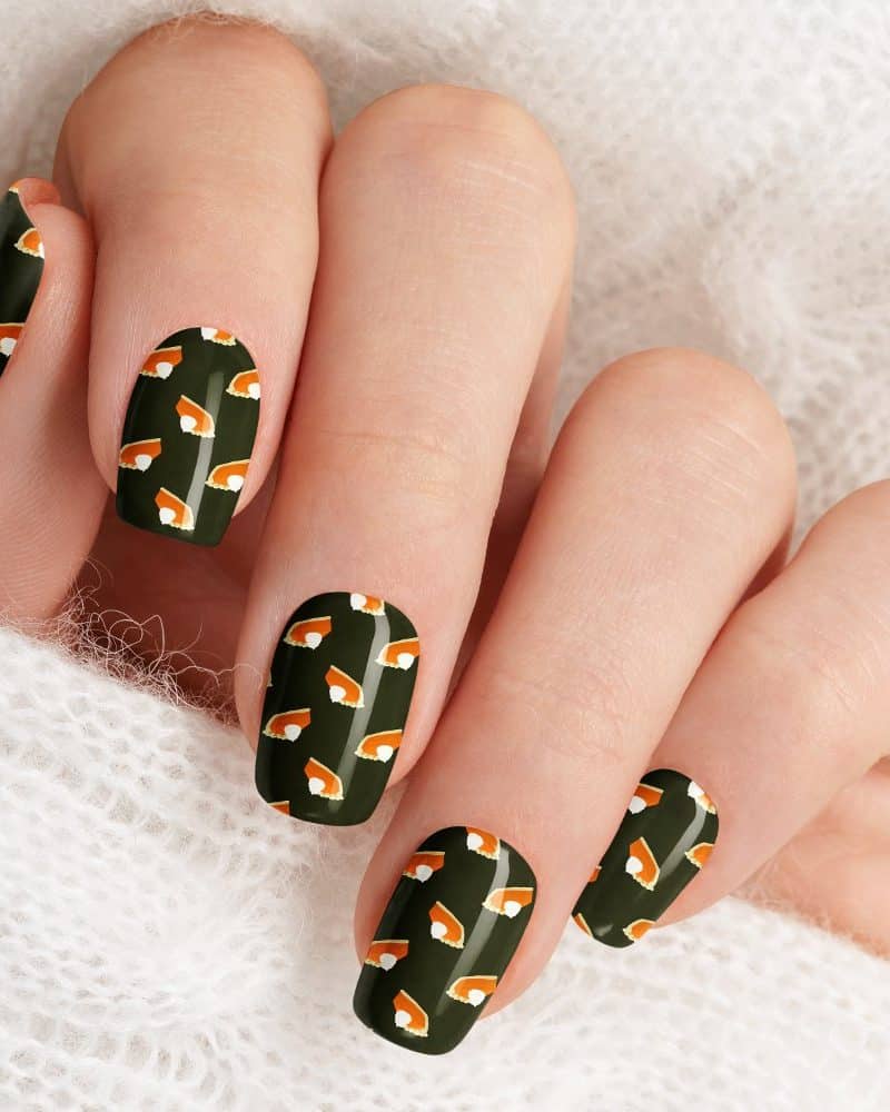 Close-up of a hand with dark green nails decorated with a pattern of miniature pumpkin pies.