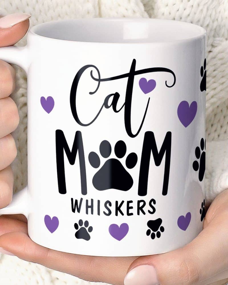 A white coffee mug with the words Cat Mom Whiskers, decorated with purple hearts and black paw prints.