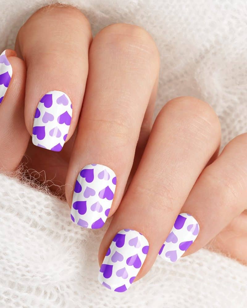 Close-up of a hand with fingernails painted with a pattern of purple hearts on a white background.